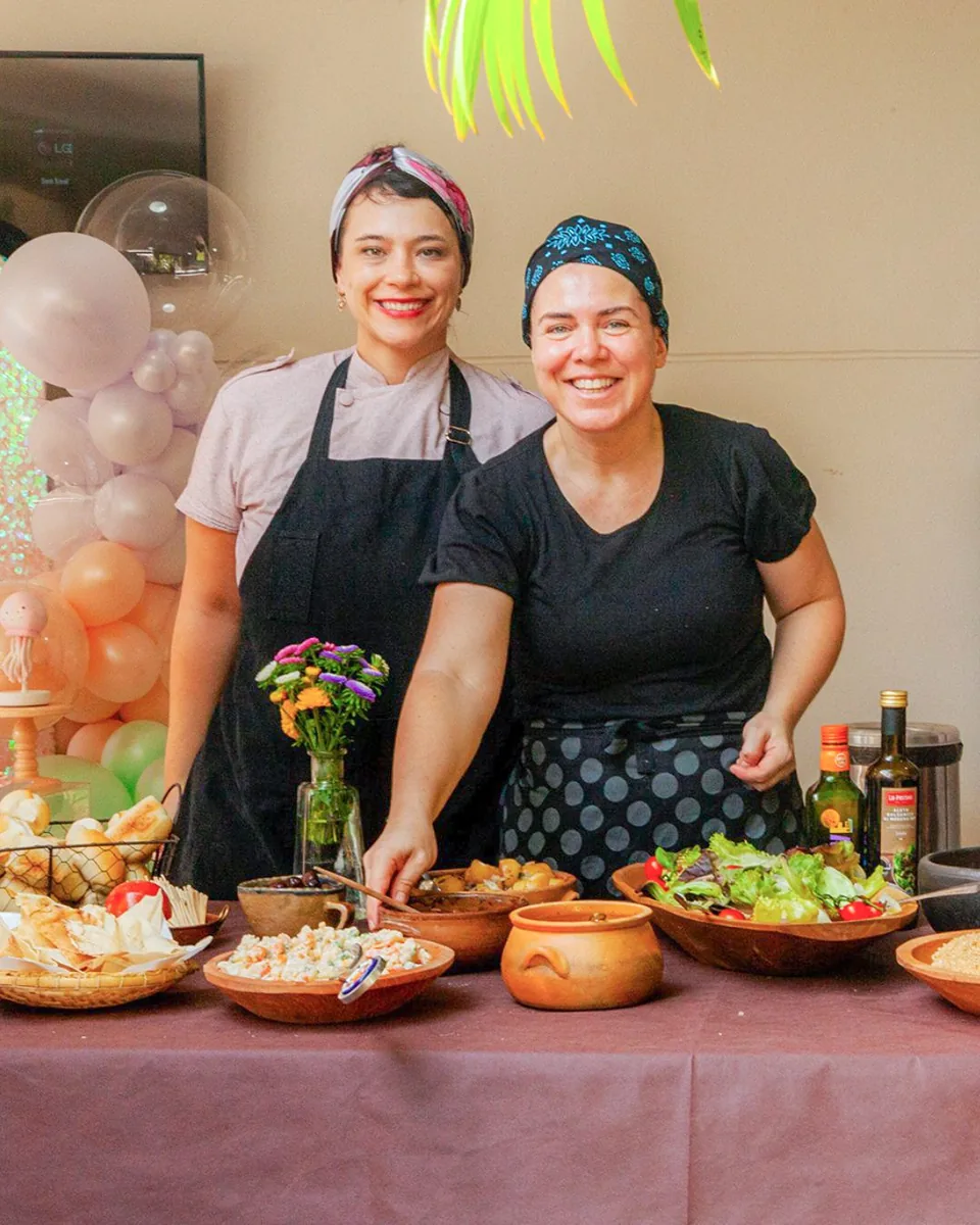 Dani Lisboa em bastidor de evento, ao lado de outra chef, com buffet montado